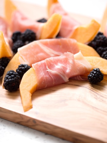 Cantaloupe slices wrapped in prosciutto with blackberries on a cutting board