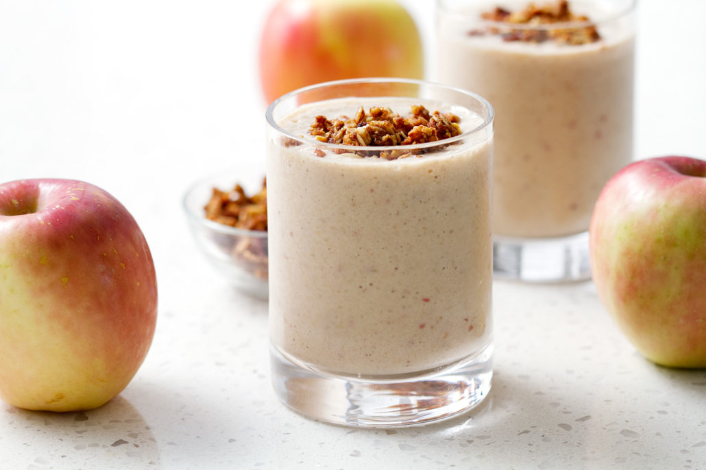 glass of smoothie surrounded by apples on white background