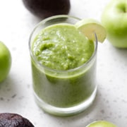 glass of green smoothie surrounded by apples and avocados on white background