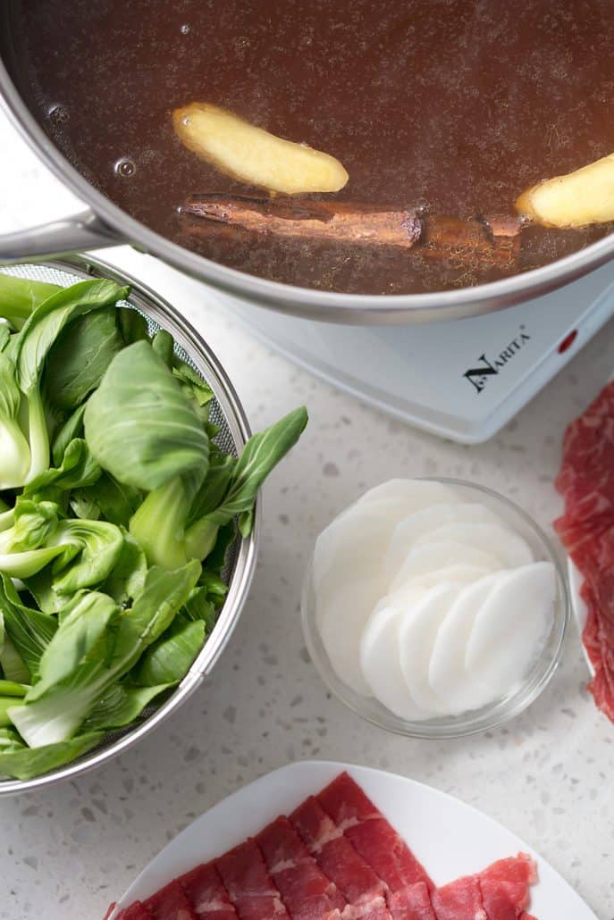 bok choy, daikon and raw meat around hot pot with cinnamon stick and slices of ginger