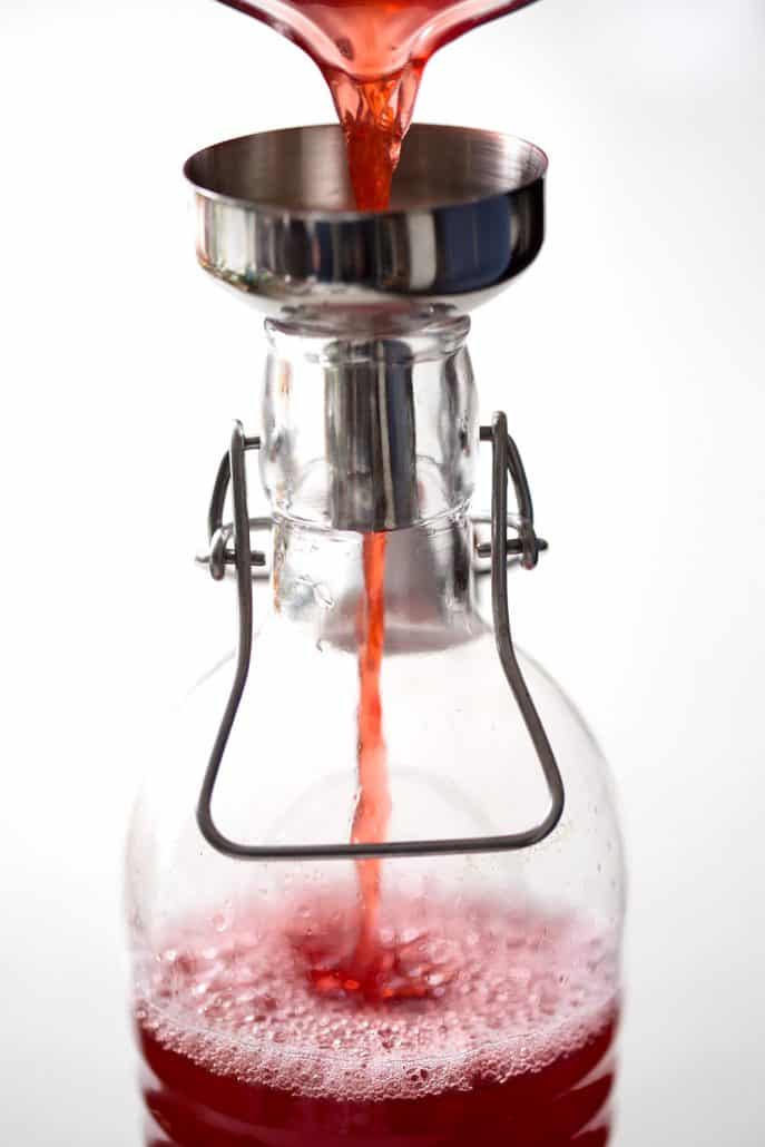 pink lombucha being poured into bottle through funnel on white background