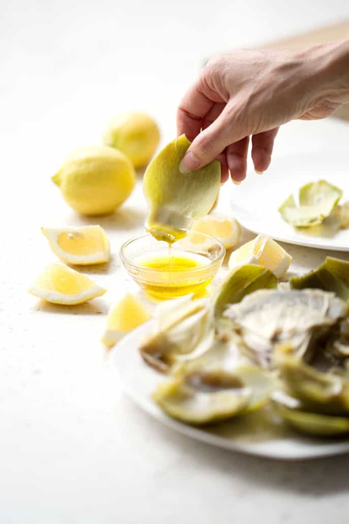 dipping artichoke leaf into butter dish on white counter