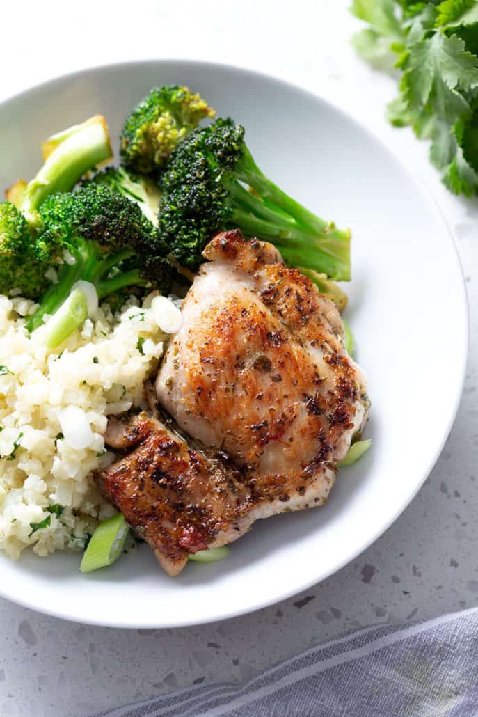 Chicken thigh resting on rice and broccoli on white bowl