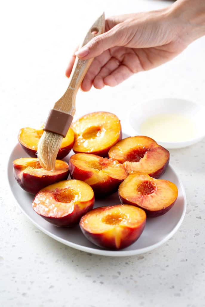 fresh peach halves being brushed with oil on platter on white counter