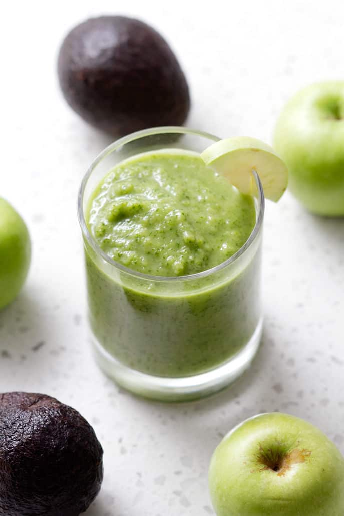 green smoothie in a glass surrounded by avocados and green apples