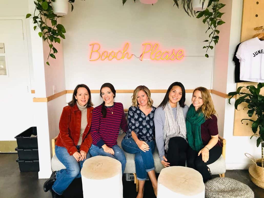 group of women sitting on couch under neon sign that says booch please