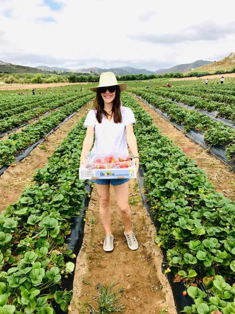 woman in hat and sunglasses posing with flat of strawberries in field