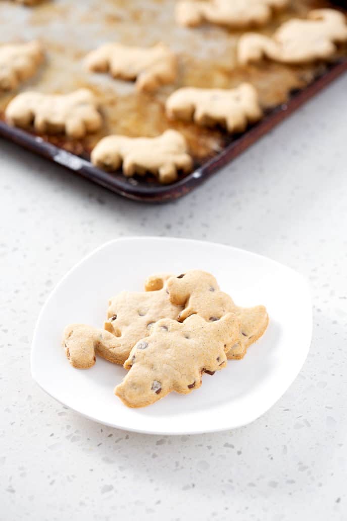 dinosaur cut out cookies on white plate with more cookies on baking sheet