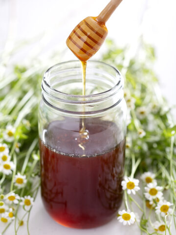 honey dipper drizzling honey into a jar with bee surrounded by flowers
