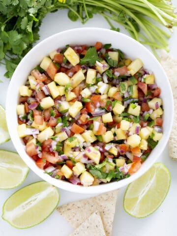 bowl of pineapple pico de gallo surrounded by cilantro, limes, tortilla chips