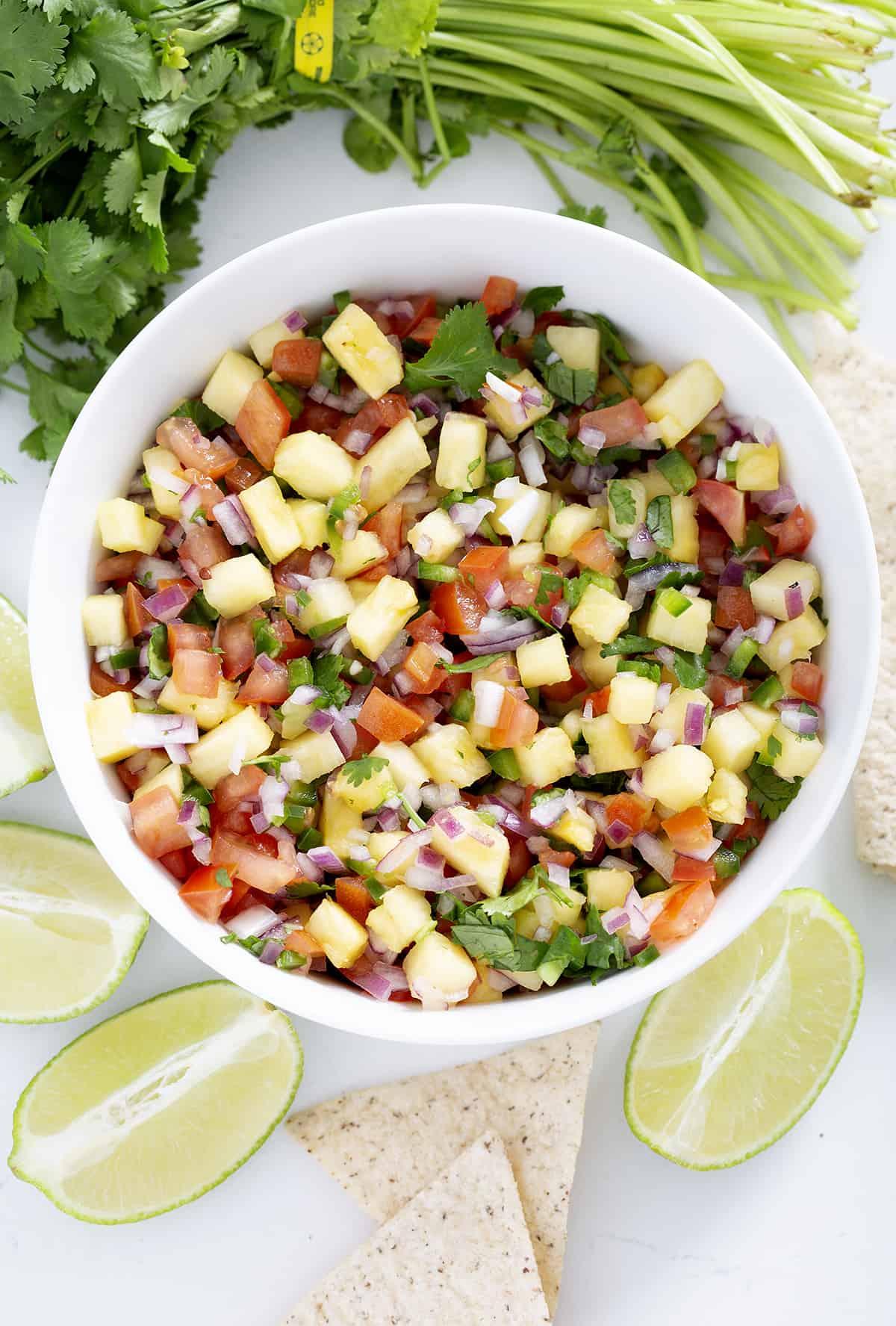 bowl of pineapple pico de gallo surrounded by limes, cilantro, tortilla chips