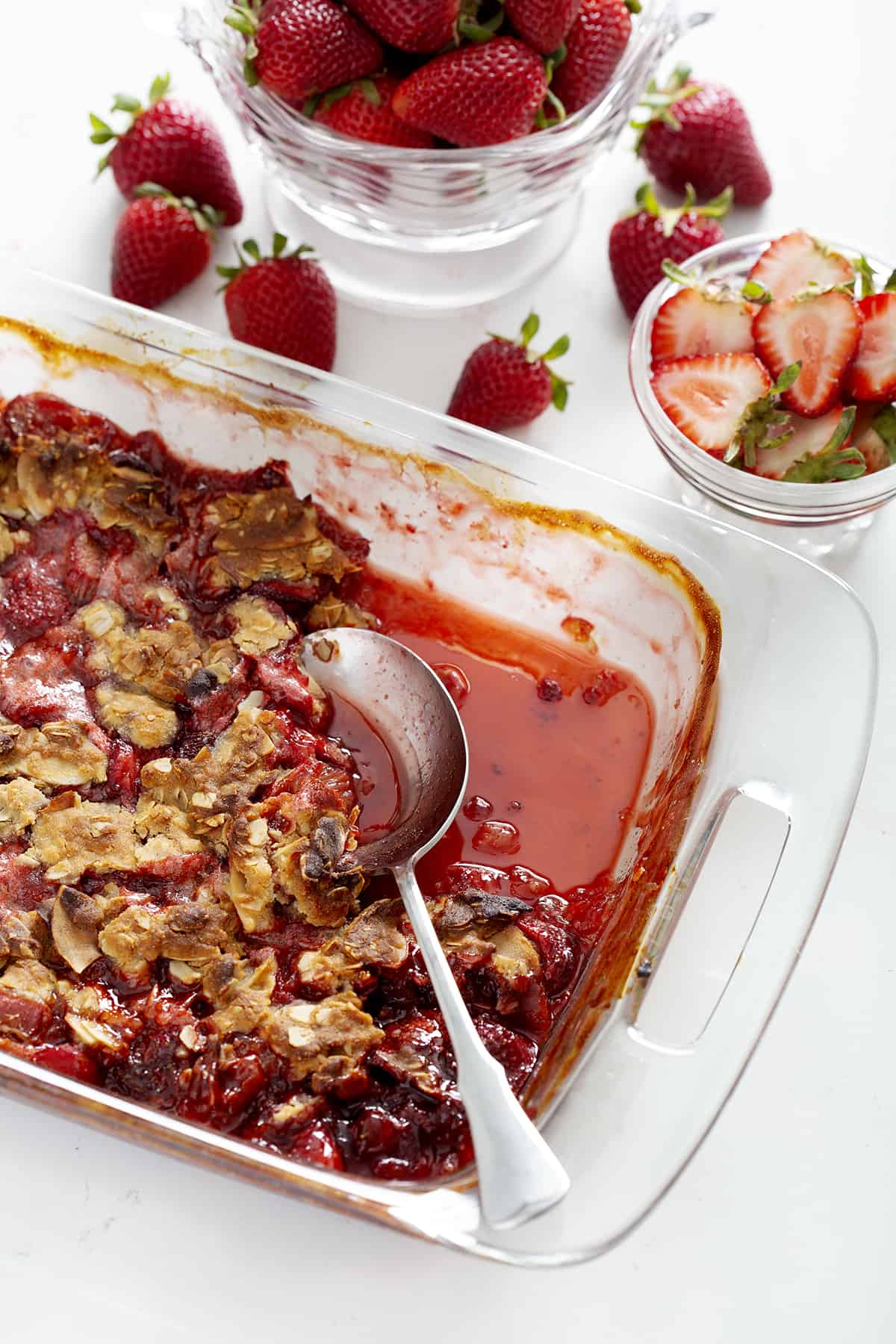 glass pan of strawberry rhubarb crumble with strawberries and serving spoon