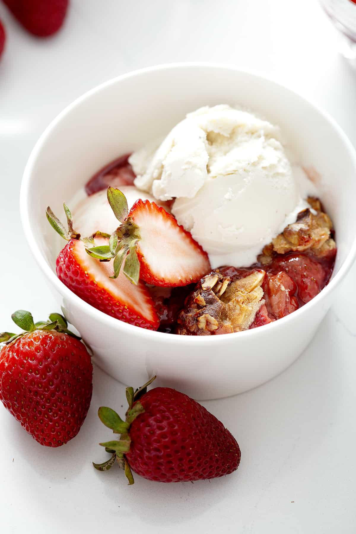 white bowl of strawberry rhubarb crumble with ice cream scoop and strawberries