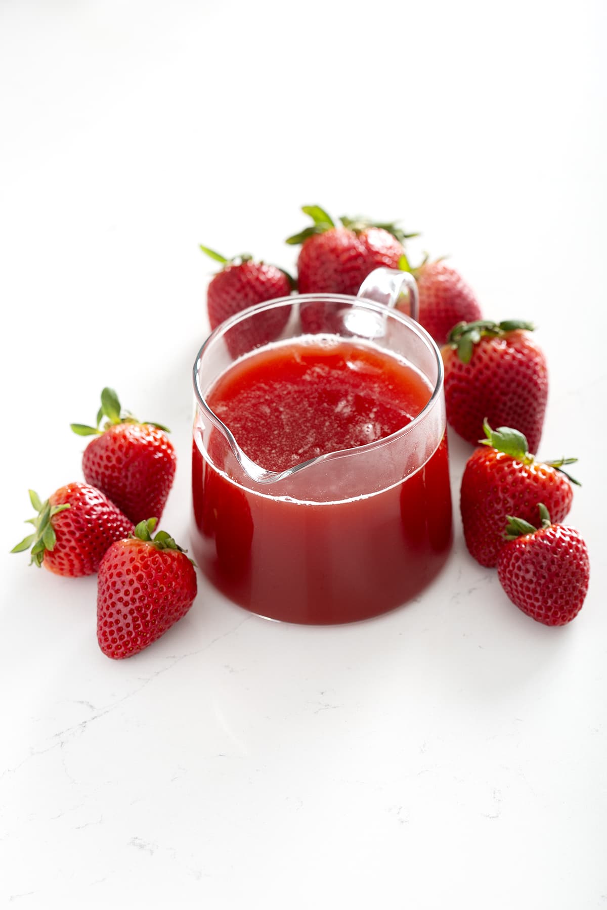 small pitcher of red Strawberry Rhubarb Syrup surrounded by strawberries